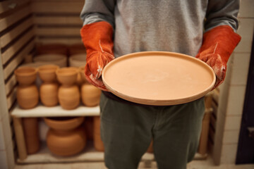 Ceramist standing with a clay plate by a kiln in his workshop
