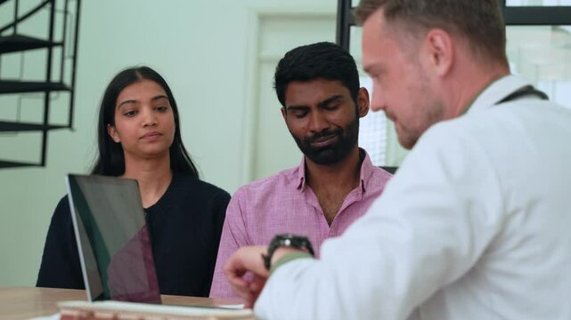 Caucasian doctor showing information on his smartwatch to an indian couple, possibly explaining diagnosis or treatment options during a consultation in a hospital room