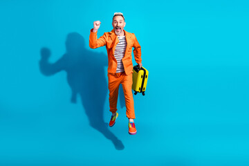 Stylish mature man in an orange suit holding a yellow suitcase, enthusiastic pose on a vibrant blue...