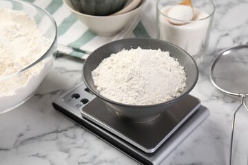 Digital kitchen scale with bowl of flour and other products on white marble table, closeup