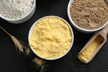 Different types of flour in bowls, scoop and spike on black textured table, flat lay