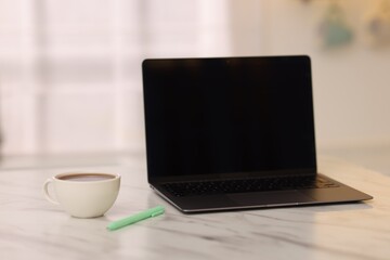Laptop with blank screen, cup of hot drink and pen on white marble table in kitchen. Mockup for design