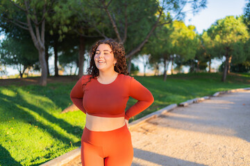 Happy plus size sportswoman wearing orange sportswear standing in a park with hands on hips, smiling and enjoying her workout