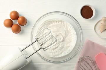 Bowl with whipped cream, hand mixer and ingredients on white wooden table, flat lay