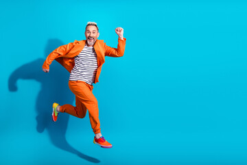 Charismatic mature man dressed stylishly in an orange suit leaping joyfully against a vibrant blue...