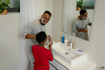 African American father teaching son to shave in bathroom, sharing joyful bonding moment