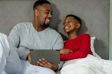 African American father and son smiling while watching tablet together in cozy bedroom
