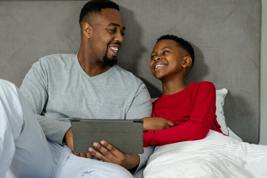 African American father and son smiling while watching tablet together in cozy bedroom