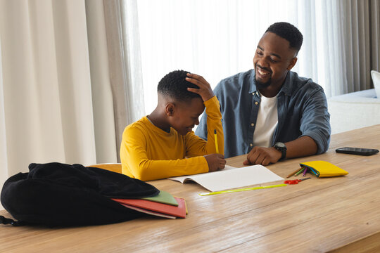 African American father and son collaborating on homework at wooden dining table indoors with pens