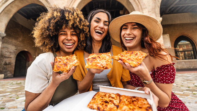 Fototapeta Three young female friends eating pizza sitting outside - Multiracial women enjoying street food in the city - Italian food culture and summer vacations concept