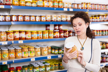 Female shopper selects glass jar of sauerkraut at a grocery store. Woman shopper reading expiration date on product label