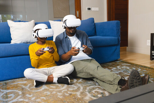 African American father and son using VR headsets indoors on blue couch with gaming controllers