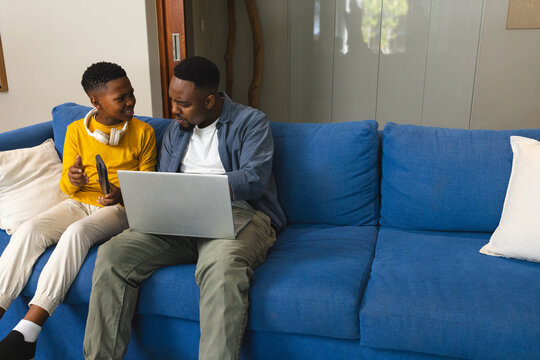 African American father and son using laptop and smartphone on blue sofa, smiling - Powered by Adobe