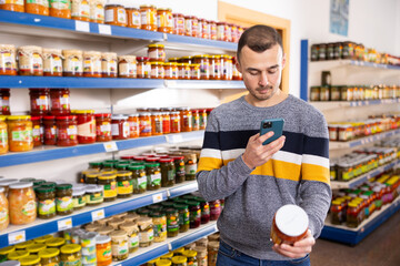 Man in store of Russian goods photographs jar canned lecho - stewed salad made from bell pepper, tomato sauce. Buyer scan QR code compares item in store and in mobile application