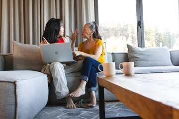 Asian mother and daughter chatting on couch at cozy living room, using laptop and white mugs