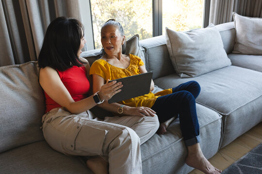 Asian young woman holding tablet while elderly woman chatting on couch at cozy living room