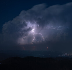 Dramatic lightning strikes burst through dense storm clouds over a dark mountainous terrain,...