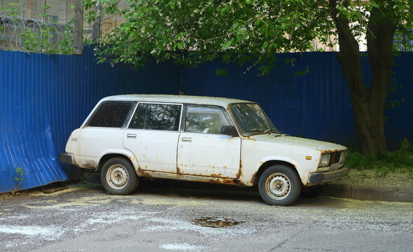 An old white rusty car is parked near a blue fence, Stakhanovtsev Street, Saint Petersburg, Russia, May 22, 2025
