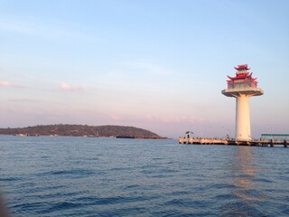 Scenic view of a lighthouse with traditional Asian architectural style on a pier at sunset, overlooking a calm sea and distant island. Peaceful maritime landscape perfect for travel or coastal themes.