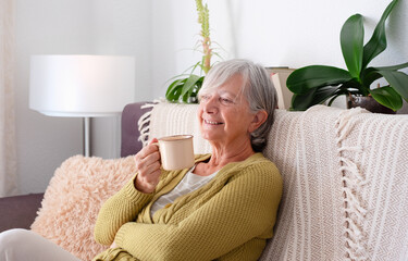 Smiling Senior Woman Sitting Indoors in Cozy Sofa having a relaxing moments enjoying a cup of coffee. Elderly Lady and retirement lifestyle