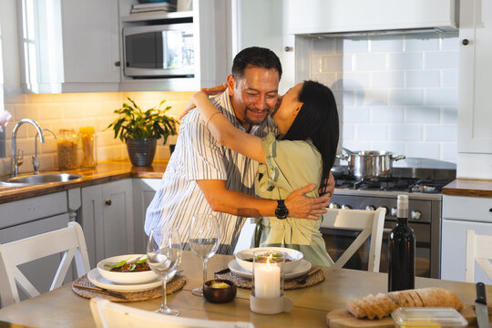 Asian couple embracing affectionately in modern kitchen with plates, wine glasses and candle