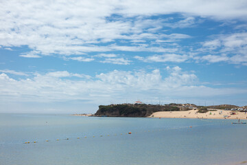 A scenic view of the beach in Vila Nova de Milfontes, Alentejo, Portugal, with calm waters, soft sand, and a cliff in the background under a vibrant blue sky with scattered clouds.
