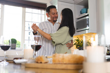 Asian couple dancing in modern kitchen, sharing intimate moment with wine glasses and candles
