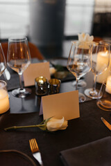 Elegant table setting for a sophisticated celebration on a luxurious black table. Golden candleholders, wine glasses, white rose, napkins, and a paper name card create a refined atmosphere.