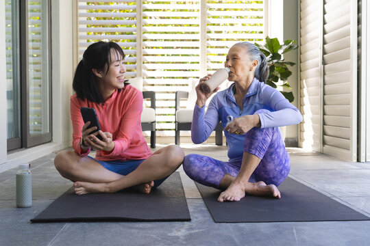 Sitting Asian mother and daughter holding smartphone and water bottle on veranda, with yoga mats - Powered by Adobe