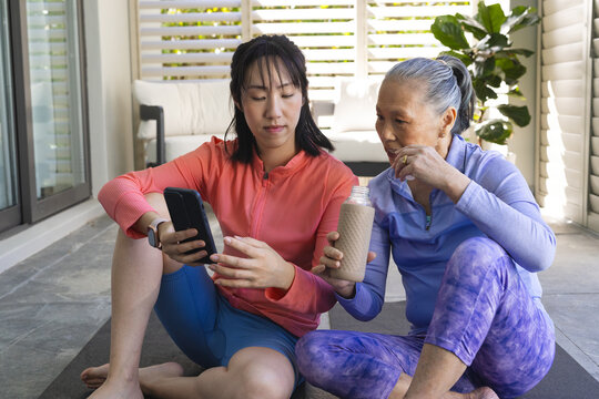 Practicing yoga Asian mother and daughter on yoga mats on patio, with smartphone and drink bottle - Powered by Adobe