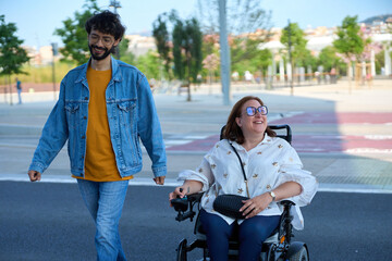 Young man walking with disabled woman in wheelchair in a city park - wheelchair and disability...