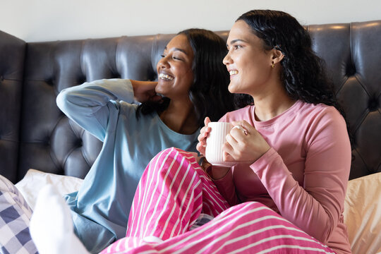 Relaxing diverse friends reclining on bed, holding white ceramic mug by dark tufted headboard
