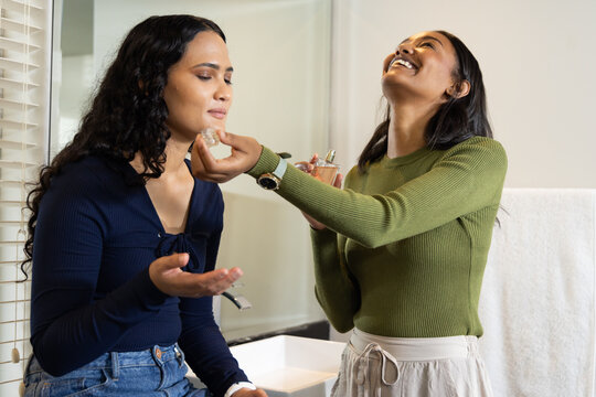 Diverse female friends applying perfume in bathroom, with white sink, towel rack and window blinds