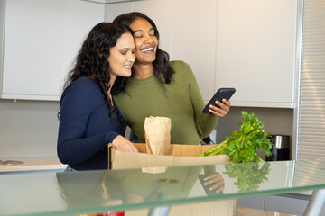 Unpacking groceries Diverse female friends checking smartphone on kitchen island with celery