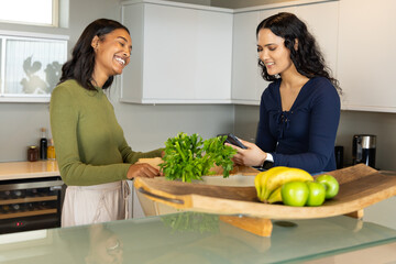 Diverse female friends unpacking groceries in home kitchen, with paper bag parsley and smartphone