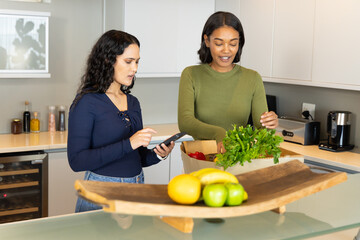 Using tablet diverse female friends unpacking at kitchen island, with paper bag and fruit tray