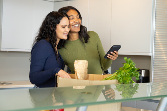 Unpacking groceries Diverse female friends checking smartphone on kitchen island with celery