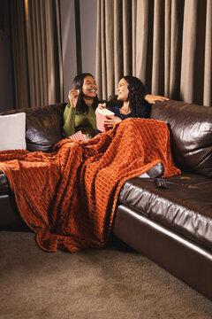 Laughing diverse female friends sharing popcorn on leather sofa in living room, with blanket