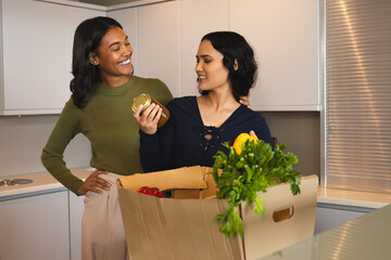 Unpacking groceries diverse female friends at kitchen island, with cardboard box of fresh produce