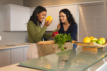 Diverse female friends unpacking produce box on kitchen island, with bell peppers and fruit bowl