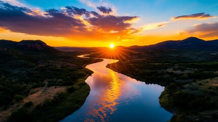 River flows through a valley landscape as the sun sets behind distant mountains, creating reflections in the water.