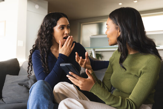 Holding smartphone Diverse female friends sitting on grey couch in modern living room, with kitchen - Powered by Adobe