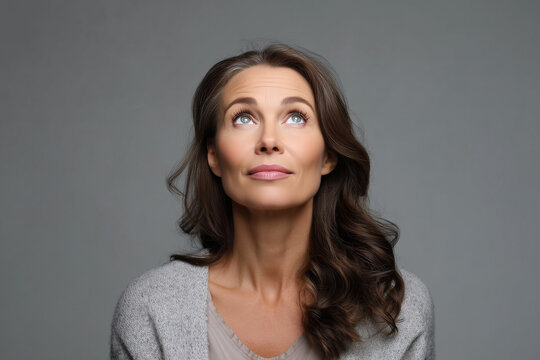 mid age beautiful woman looking upwards on a gray background, she is looking questioned and confused
