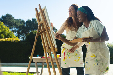 Painting diverse female friends collaborating at easel on patio, holding palette and paintbrushes