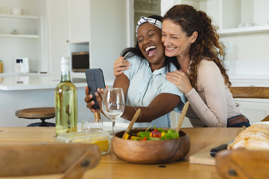 Laughing Diverse female friends embracing as viewing smartphone at kitchen table, with salad bowl