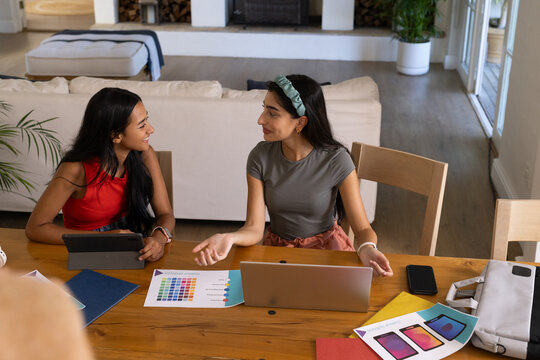 Diverse female friends chatting at table in living area, with laptop and color charts