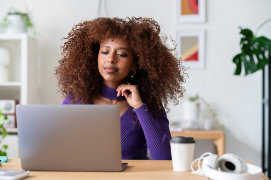 A woman with vibrant curly hair is intently focused on her laptop screen, appearing to be working or studying diligently from her home office setup. She seems engaged in her task.