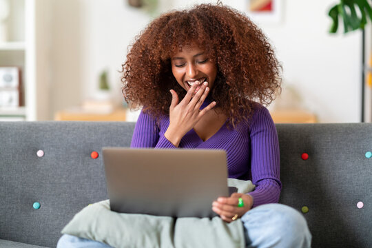 A woman with curly hair sits on a couch, looking at her laptop. She covers her mouth, reacting to something she sees on the screen, perhaps finding it funny or surprising.