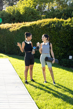Chatting diverse female friends on boardwalk in sunny garden, holding yoga mats and water bottles