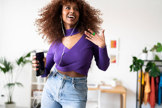 A joyful woman with curly hair is dancing and listening to music through wired headphones connected to her phone, expressing happiness and freedom in her home.